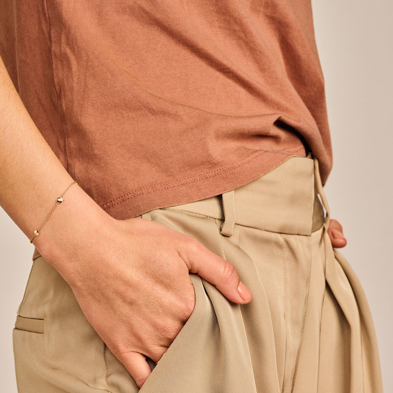 Fijne armband van 14k goud met subtiele schakels om pols bij beige broek en bruin shirt.